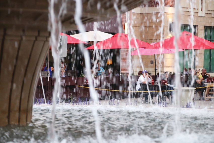 Patrons eating outdoors are seen through water falling from The Streets at SouthGlenn’s fountain, where small numbers of people also gathered.