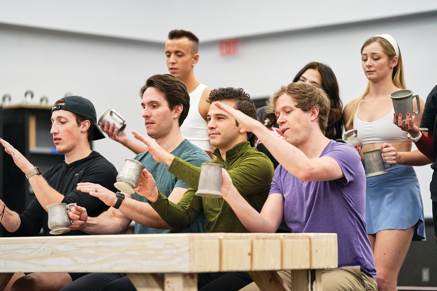 Kevin Hack (Gaston) and Nicolas Garza (Lefou) sit center on the bench surrounded by the ensemble during a pub scene.
